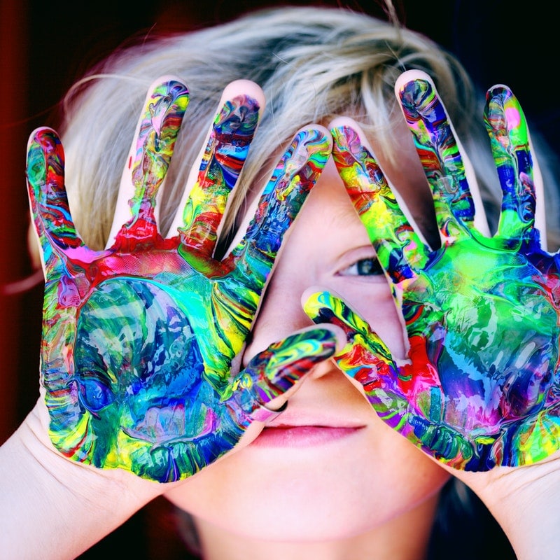 boy with different coloured paints all over his hands. Peeking through his hands at the camera