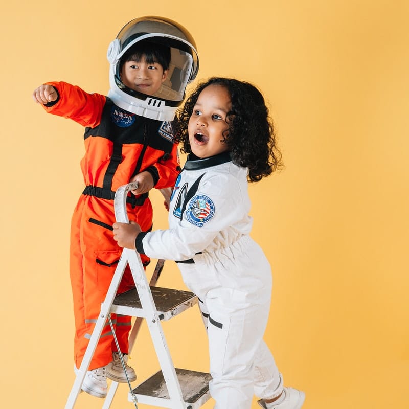 Kids using their gross motor skills to climb a ladder.  Both kids are dressed as astronauts; the boy is on top of the ladder pointing to something off in the distance