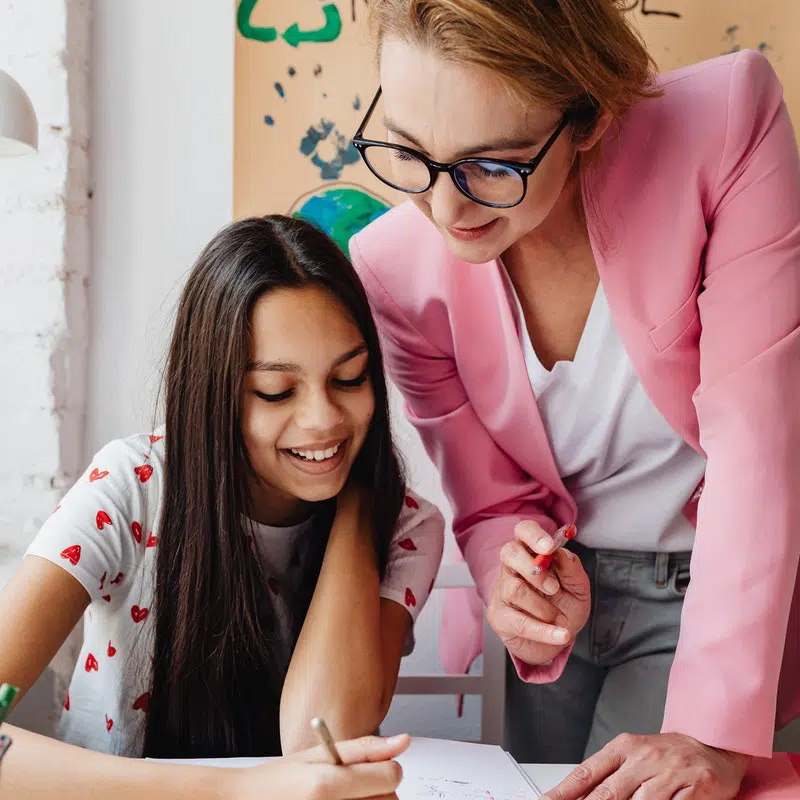 Child learning Organization Skills from teacher. Student is smiling and teacher seems pleased. They are both looking at her work.