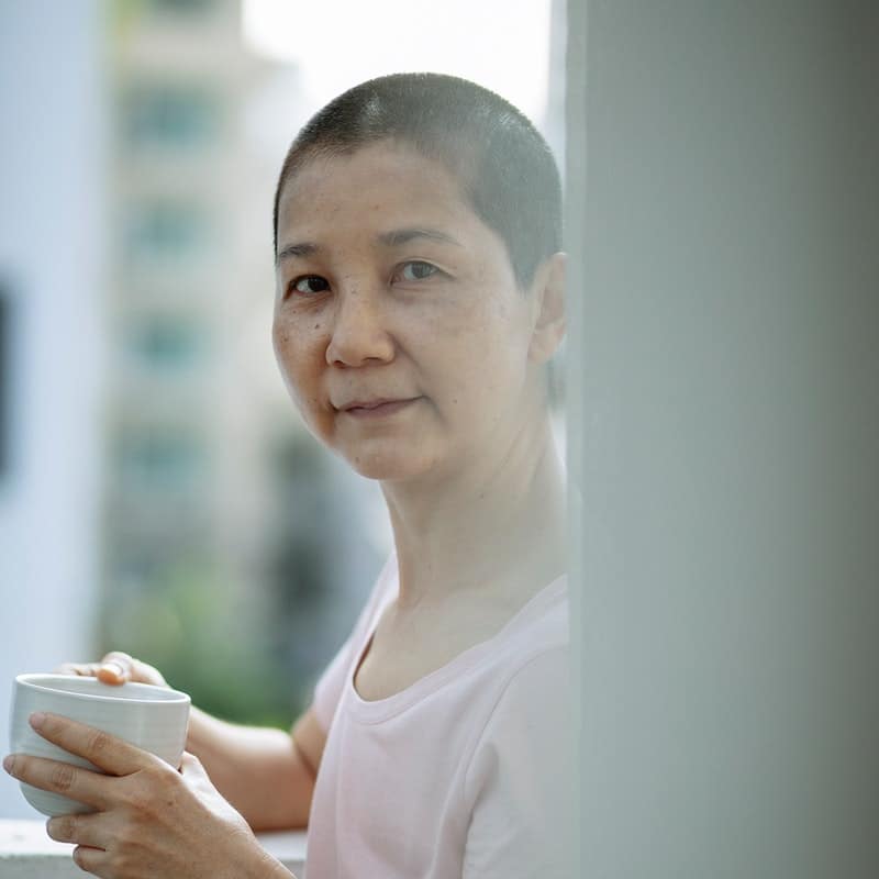 Cancer patient about to drink coffee, looking at the camera.  She is on a balcony of a building.
