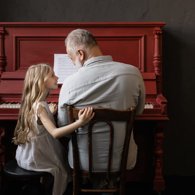 Senior playing music on a red piano with granddaughter looking on